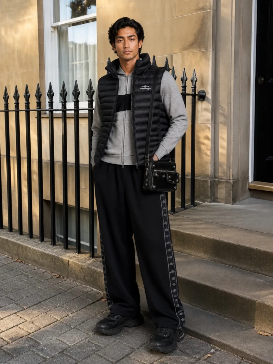 Balenciaga’s quilted gilet sits over a light grey technical hoodie and fluid black logo track pants for a monochrome, city-sport uniform. Chunky black runners echo the padded volumes while the compact moto crossbody brings a hit of hardware and texture without disturbing the clean lines. It is a quietly assertive streetwear set built for movement, from airport concourses to late-night walks home.