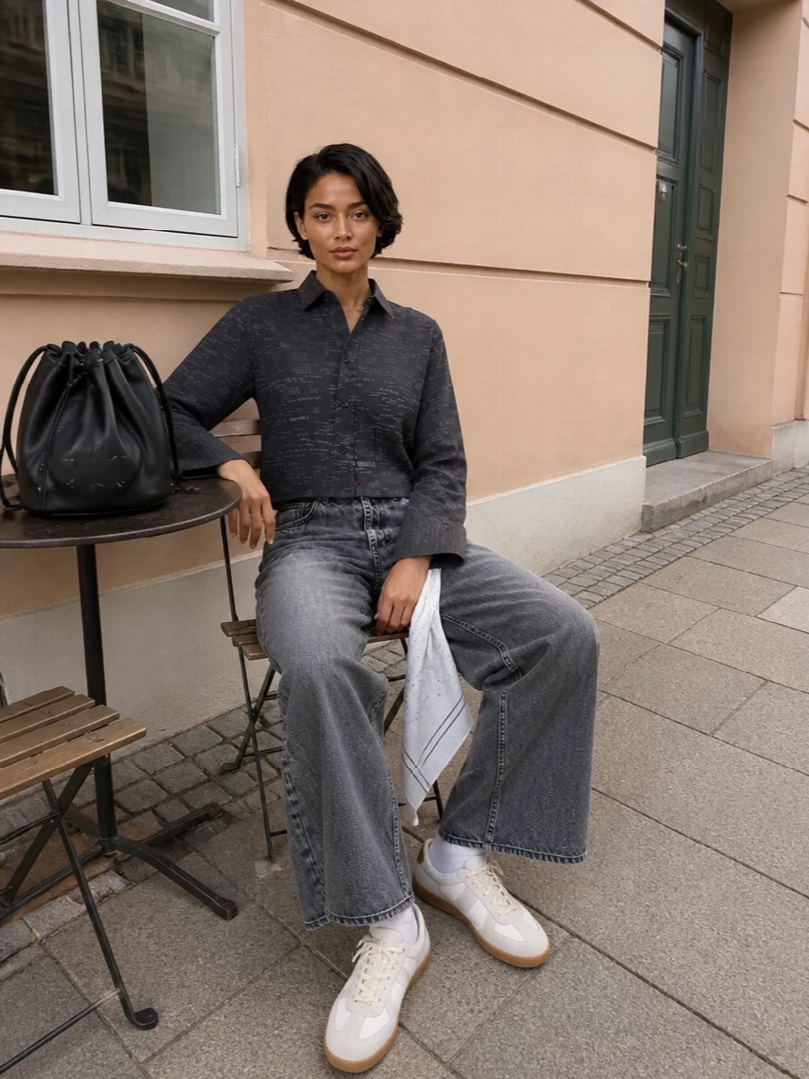 Washed charcoal denim meets Margiela’s cropped printed shirt for a clean, graphic take on wide-leg streetwear. Pale grey sneakers and a black bucket bag keep the mood precise but relaxed, while a soft silk square lifts the darker palette just enough. The proportions stay long and lean, with the high rise and cropped top creating an easy, modern column through the leg.