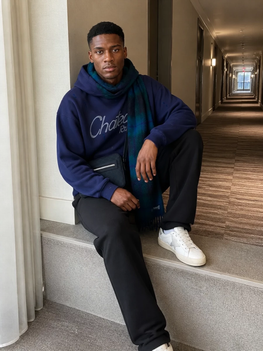 Gallery Dept. on top and bottom keeps this relaxed navy hoodie and washed-black track pant combo feeling like a single, intentional set instead of just sweats. Soft neutrals at the sneakers and a compact navy crossbody keep the line clean, while the moody blue-green scarf pulls out the hoodie’s splatter tones for a cold-weather street uniform that feels easy but considered.