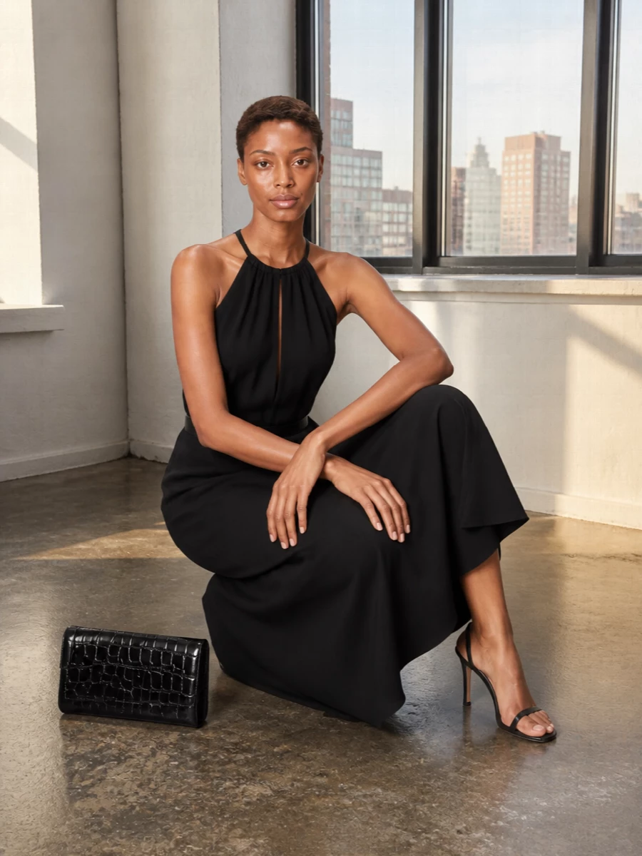 A fluid black halter maxi is cinched lightly at the waist, so the volume feels intentional rather than overwhelming. Bare black stilettos and a structured croc clutch keep everything sleek and city-focused, letting the open back read as the main detail. The result is quietly assured evening dressing that moves beautifully but never feels overworked.