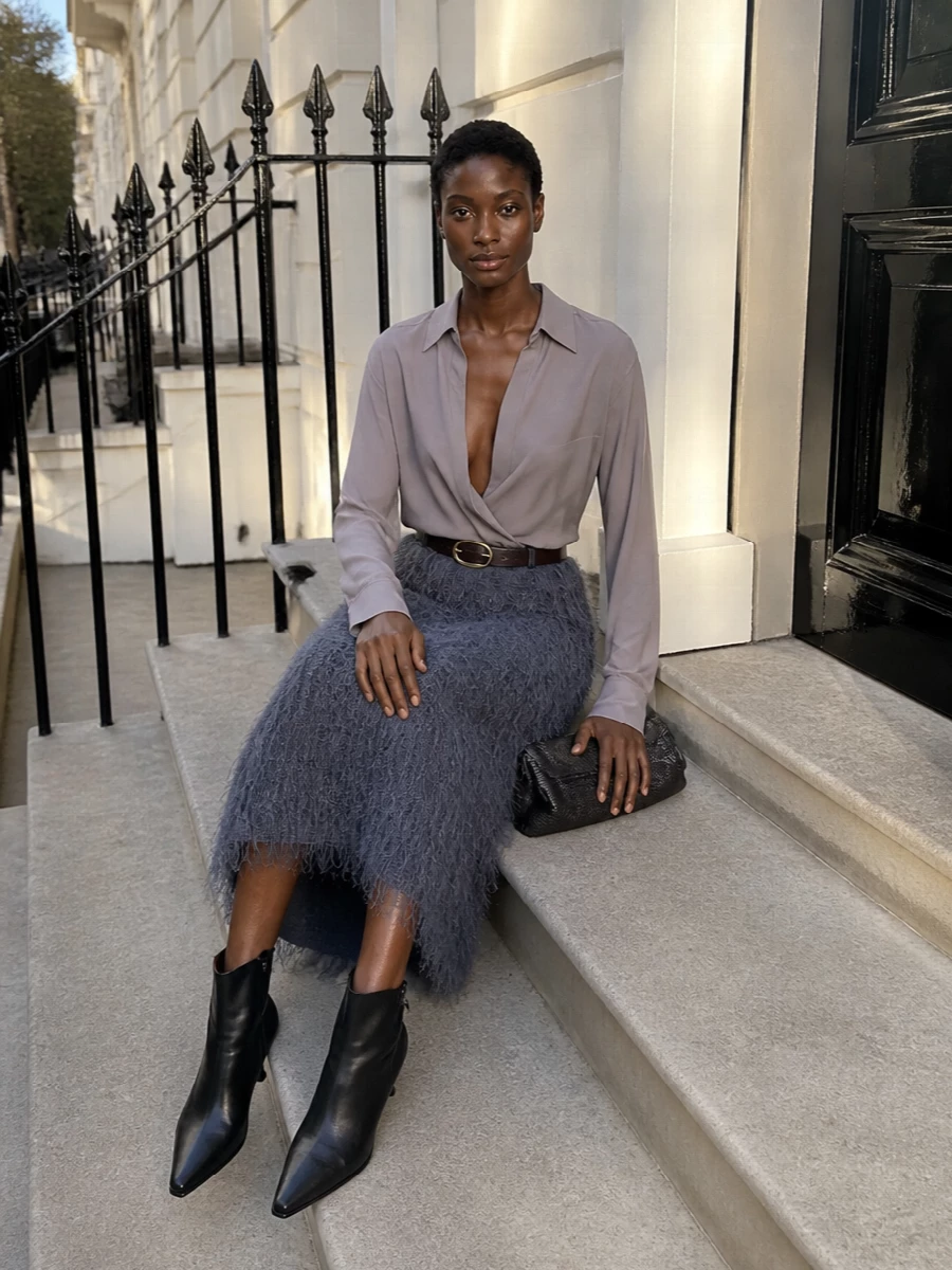 A slate grey fringe midi meets a muted mauve wrap top for a softly sculpted column that feels polished yet quietly dramatic. Black sculpted ankle boots and a gunmetal clutch sharpen the line, keeping the focus on texture and movement. A dark chocolate belt offers the option to cinch the waist and further refine the evening silhouette.