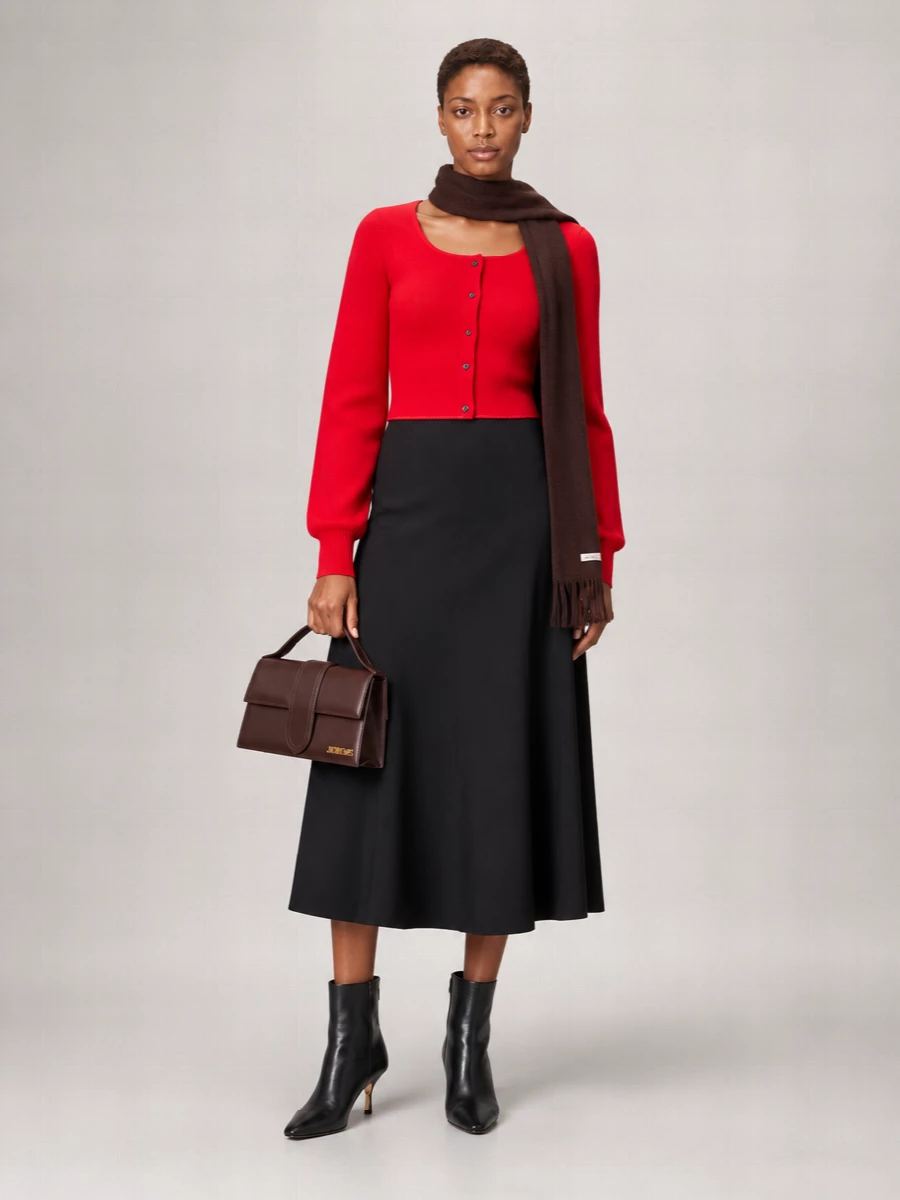 A vivid red Jacquemus cardigan becomes the focal point against a pure black high-waisted flared midi, keeping the silhouette long, clean and quietly assertive. Sculpted black ankle boots sharpen the line, while a chocolate top-handle bag and matching scarf introduce just enough depth for a warm, modern city ensemble. The result is minimalist and polished, with color used deliberately rather than loudly.