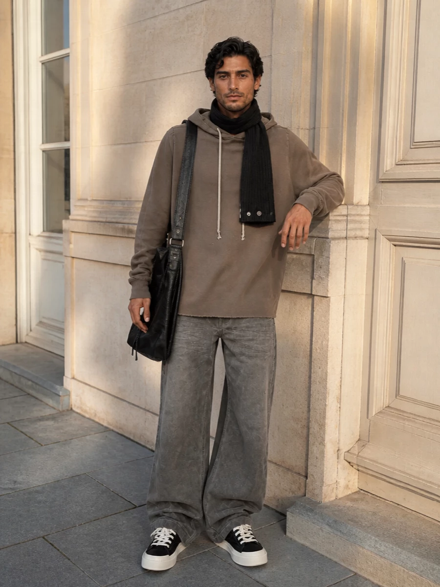 Wide taupe-grey denim grounds a washed taupe longline hoodie for an all-Rick, shadowy neutral story that feels easy but intentional. Chunky black and eggshell high-tops and a slouched black leather messenger add weight and contrast while keeping everything street facing. A snapped ribbed scarf runs the line between utility and drama, finishing the silhouette with that subtle Rick hardware detail.