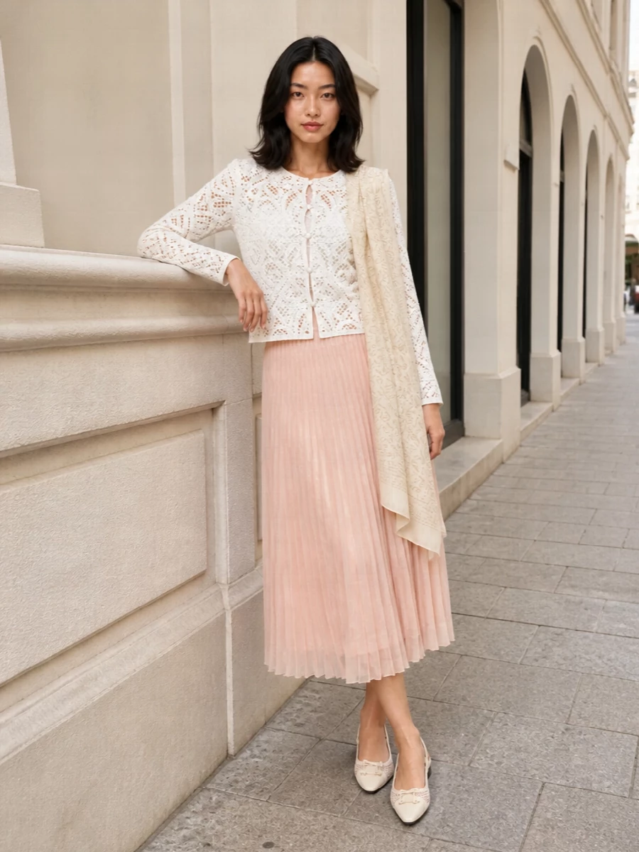 A soft ivory lace cardigan tops a blush nude pleated midi, creating a light, romantic column of texture that feels polished without stiffness. Ivory mesh slingbacks and a buttercream structured shoulder bag keep the palette calm and tonal, letting the mix of lace and pleats stay the quiet focal point. The paisley stole slips in as a barely-there pattern, adding depth while preserving the minimalist, quiet-luxury mood.