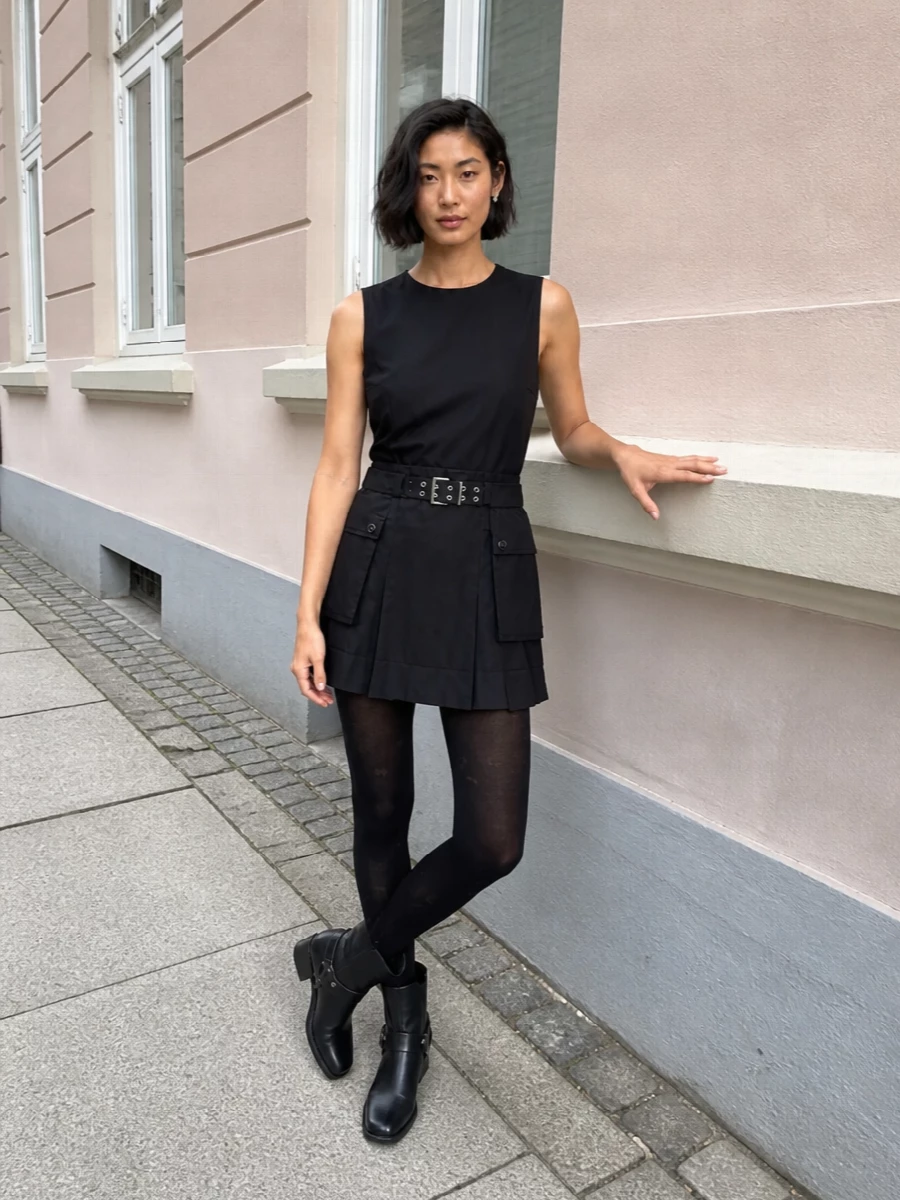 A black-on-black city set that lets the mini’s utility details do the talking. Harnessed ankle boots and sheer-ish tights ground the shorter hem, while the sculpted shoulder bag and silver orb studs keep the hardware story sharp but controlled. Head-to-toe, it reads like easy, pulled-together attitude for all-day urban pacing and after-dark detours.