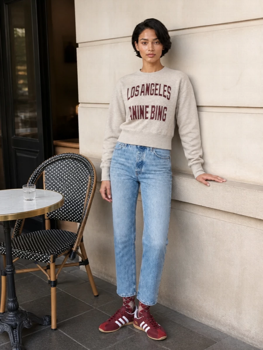 Anine Bing’s cropped collegiate fleece meets light-wash straight denim for a relaxed LA uniform that feels easy but thought through. Burgundy Adidas sneakers and matching argyle socks tie back to the graphic lettering, so the color story feels deliberate rather than random. A compact charcoal top-handle bag keeps everything grounded and city ready without fighting the sport energy.