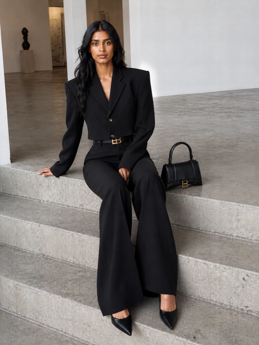 Balenciaga tailoring pushed into night mode: a cropped, sharp-shouldered jacket slicing over high-waisted black flares for a long, graphic column of black. A razor-clean pump and sculpted mini top-handle keep everything focused and controlled, while a slim belt catches the light just enough at the waist. The result is severe, quiet and distinctly subversive suiting for after-dark appointments and late dinners.