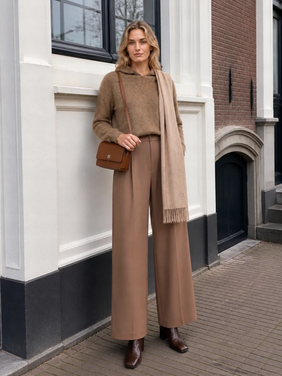 A camel hooded knit paired with fluid high-waisted wide-leg trousers creates a long, tonal column that feels relaxed yet composed. Dark, burnished ankle boots and a structured chestnut crossbody bring subtle contrast and definition, while a softly brushed beige scarf adds quiet texture and warmth. The result is a cocooning city uniform that moves easily from slow mornings to understated dinners without losing its polish.