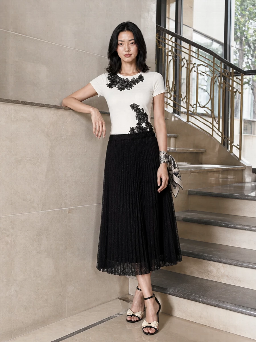 A study in monochrome romance, this pairing lets the sculpted lace tee and the sheer black pleated midi read as one fluid, evening-ready column. Black stilettos with a cream bow and a compact black shoulder bag keep the lines clean and polished, while a whisper of floral silk at the wrist or bag adds just enough softness. It is feminine, modern and quietly striking for refined dinners or cocktail gatherings.
