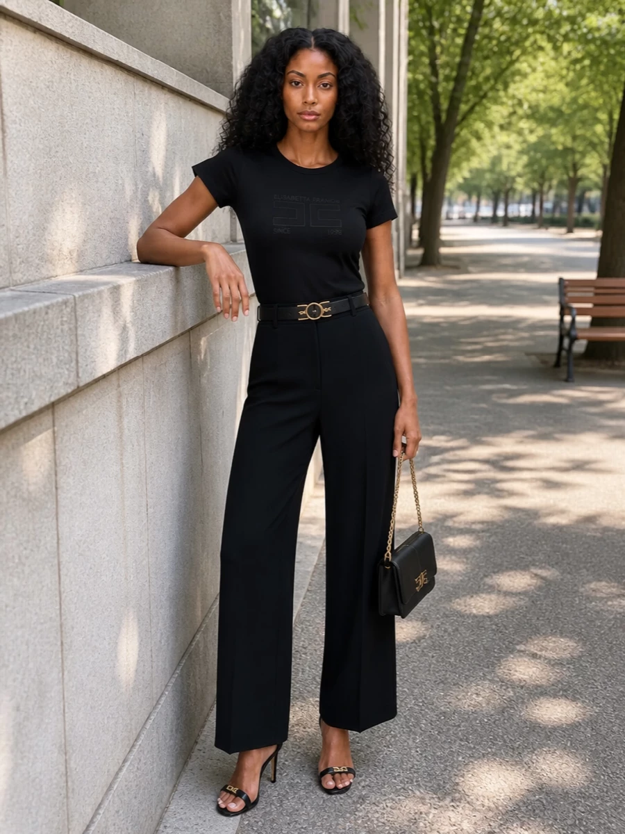 A sharp black palette keeps this outfit quietly graphic, letting the logo tee and clean tailoring feel intentional rather than casual. High-waisted wide-leg trousers and a slim belt sculpt the waist and lengthen the leg, while gold hardware on sandals and bag adds just enough shine for terrace drinks or a dressed-up city evening. It is minimal, branded and precise without ever feeling stiff.