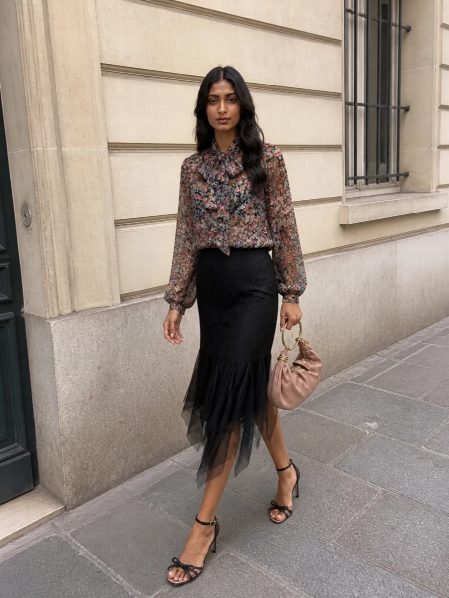 This look lets the Valentino floral pussy-bow blouse take centre stage, grounded by the dark romance of the black lace asymmetric midi skirt for a fluid, evening-ready silhouette. Nude-and-black strappy stilettos keep the line elongated and graphic, while a blush ruched clutch brings a soft touch of colour that quietly echoes the blouse’s garden palette. A sheer ivory logo scarf can be draped lightly if you want an extra whisper of couture detail without disturbing the print story.