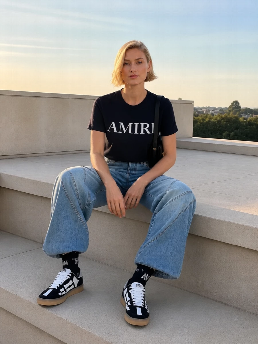 Amiri’s navy logo tee pairs with the label’s paneled wide-leg jeans for a graphic yet minimal take on streetwear, all in an easy spring-ready palette. Chunky bone-detailed sneakers ground the proportions, while a structured black shoulder bag sharpens the mood for city days that move into night. Logo socks peeking above the sneakers add a restrained, knowing nod to street styling without cluttering the silhouette.