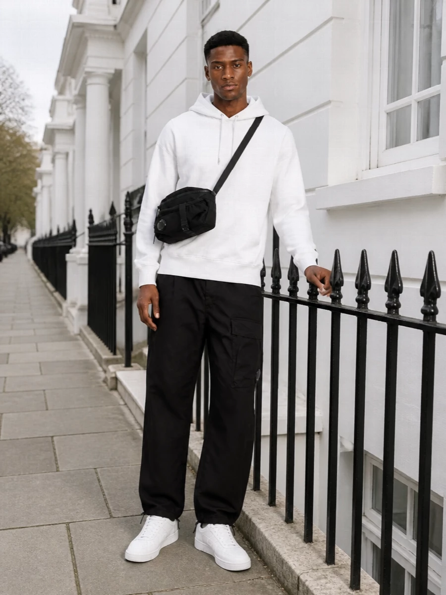 A white C.P. Company hoodie sits over crisp black technical cargos for a sharp, monochrome street-sport uniform. Clean white and grey low-top sneakers keep the lines minimal, while a compact black crossbody with lens detail ties the technical notes together for city days and travel. The result is relaxed in fit but precise in palette, easy to wear from morning coffee runs to late-night walks.