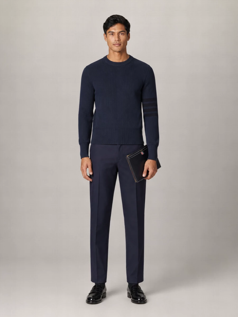 Navy on navy keeps this pairing focused and precise, with the ribbed crew sitting cleanly over straight-leg trousers for a quietly composed profile. Glossy black loafers sharpen the line without feeling stiff, while the slim navy pouch adds a disciplined finishing note that stays firmly in Thom Browne’s tailored universe.