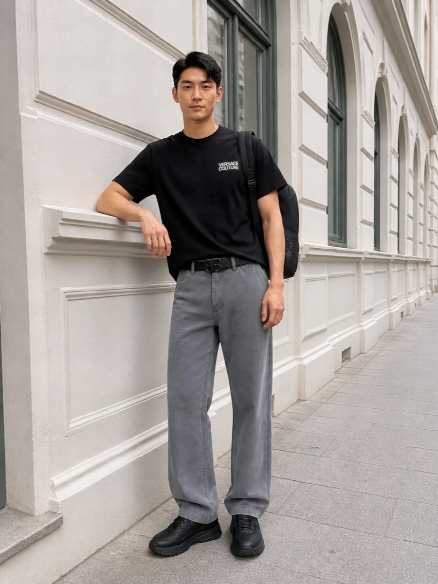 A black Versace logo tee sits over slate-grey straight-leg trousers for a monochrome streetwear uniform that feels sharp but relaxed. Chunky black sneakers and a slim dark belt ground the proportions, keeping the line clean from hip to floor. A streamlined Medusa backpack finishes it with just enough designer attitude for daily city wear.