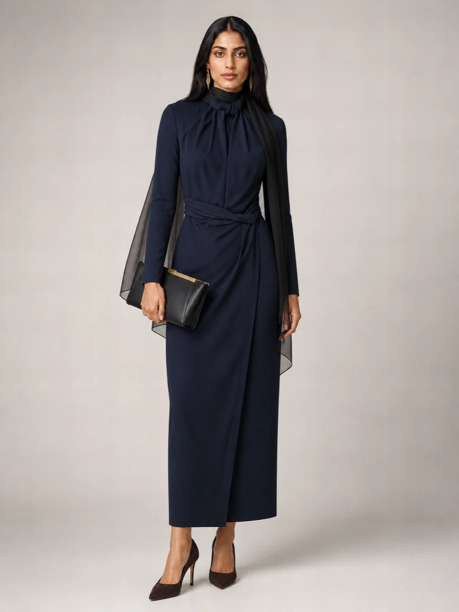 A navy wrap column dress is sharpened with deep espresso suede stilettos and a structured black clutch for a precise, cinematic evening ensemble. A single gold logo drop earring and a gauzy black wrap bring just enough light and movement to keep the silhouette quiet yet unmistakably powerful.