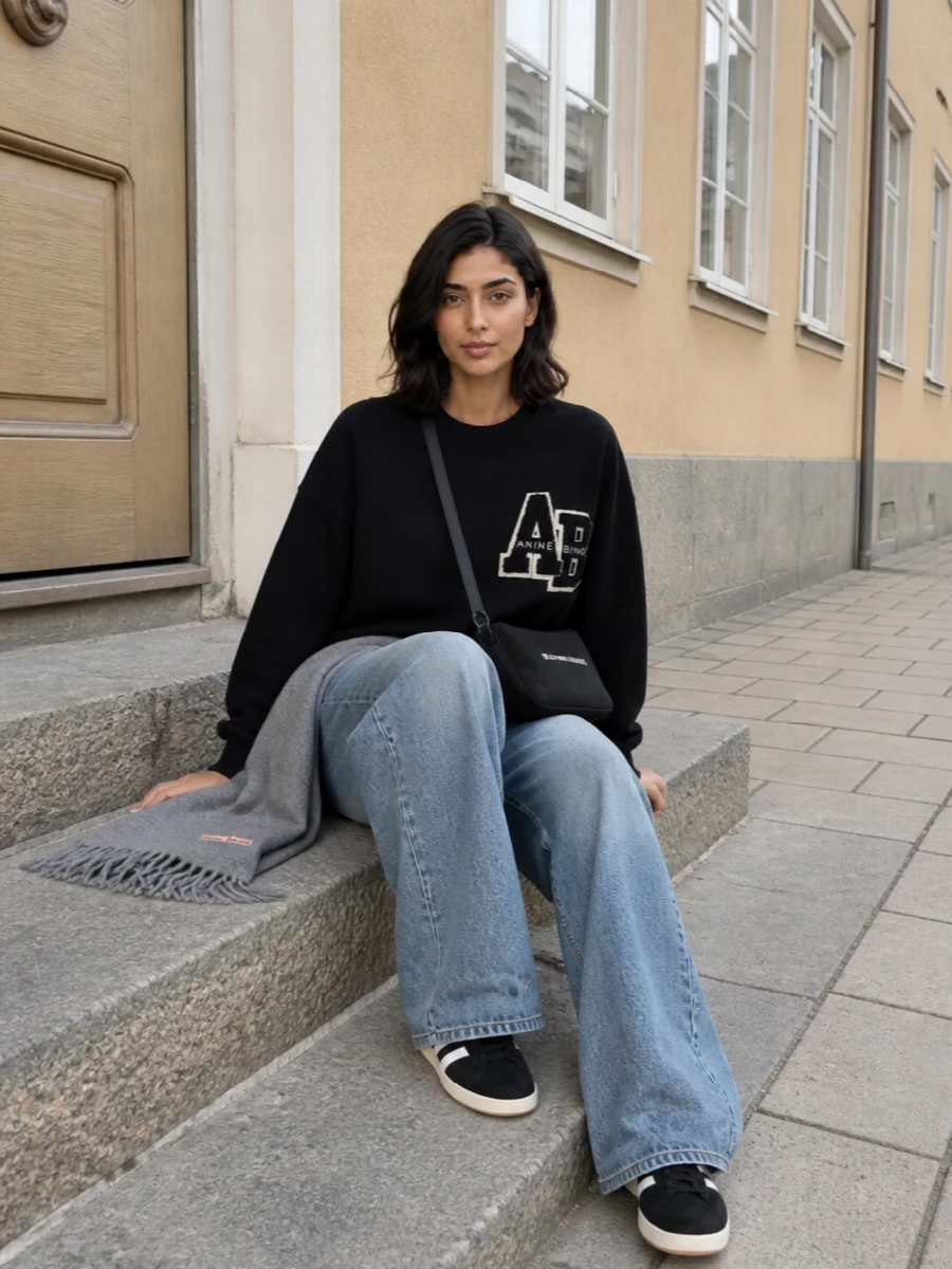 An oversized collegiate sweatshirt relaxes into high-rise wide-leg denim for a clean, slouchy streetwear uniform. Black and cream sneakers and a graphic cross-body keep the mood low-key but intentional, while a soft grey scarf adds just enough winter texture. It is an easy travel or city-errand combination that feels modern without trying too hard.