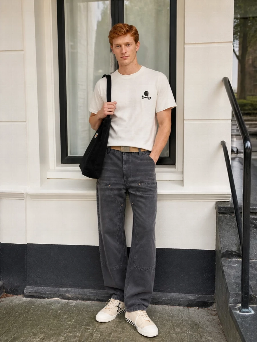 An off white graphic tee sits against washed charcoal work pants for a clean, skate-leaning contrast. Cream and black canvas sneakers echo the tee’s tones, while a black tote and sand webbing belt keep the utility story practical and tight. The result is an easy city uniform that feels considered without trying too hard.