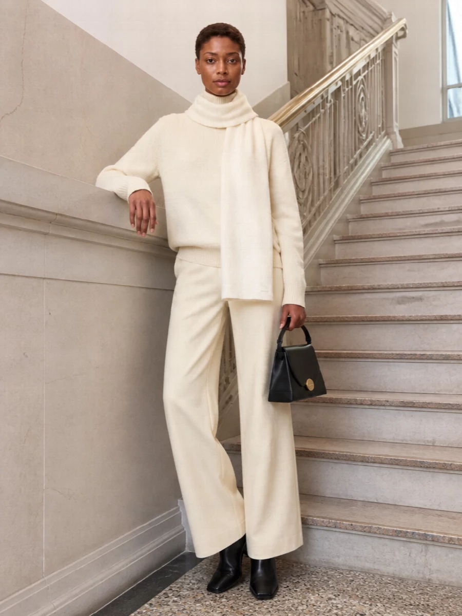 Buttercream on buttercream creates a calm column, with wide-leg tailoring giving the mock-neck knit a long, architectural line rather than weekend ease. Black sock boots and a sculpted top-handle bag cut through the softness with precise contrast, keeping the mood quietly formal. An ivory scarf adds just enough texture at the neck to feel considered in colder weather while preserving the restrained palette.