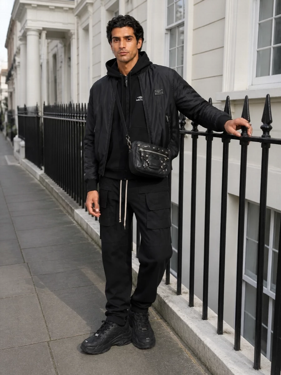 A strict black-on-black story that blends Dolce&Gabbana precision with Balenciaga and Rick Owens street attitude, this outfit reads like a considered uniform rather than loungewear. The technical bomber sits clean over a logo-forward hoodie, grounded by graphic cargos and a heavy-soled sneaker that can hold the volume. A moto-detailed crossbody adds just enough hardware to keep the monochrome from feeling flat while staying quietly aggressive and city-ready.