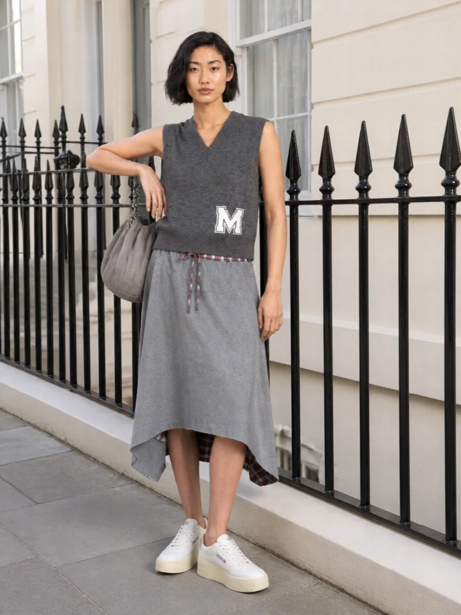 A soft grey Westwood midi meets a charcoal varsity vest for a streetwise, slightly preppy take on summer layering. Platform sneakers keep the asymmetric hem easy to move in, while a slouchy grey chain tote grounds the outfit in practical city reality. It is relaxed, graphic and ready for long days on your feet without losing attitude.