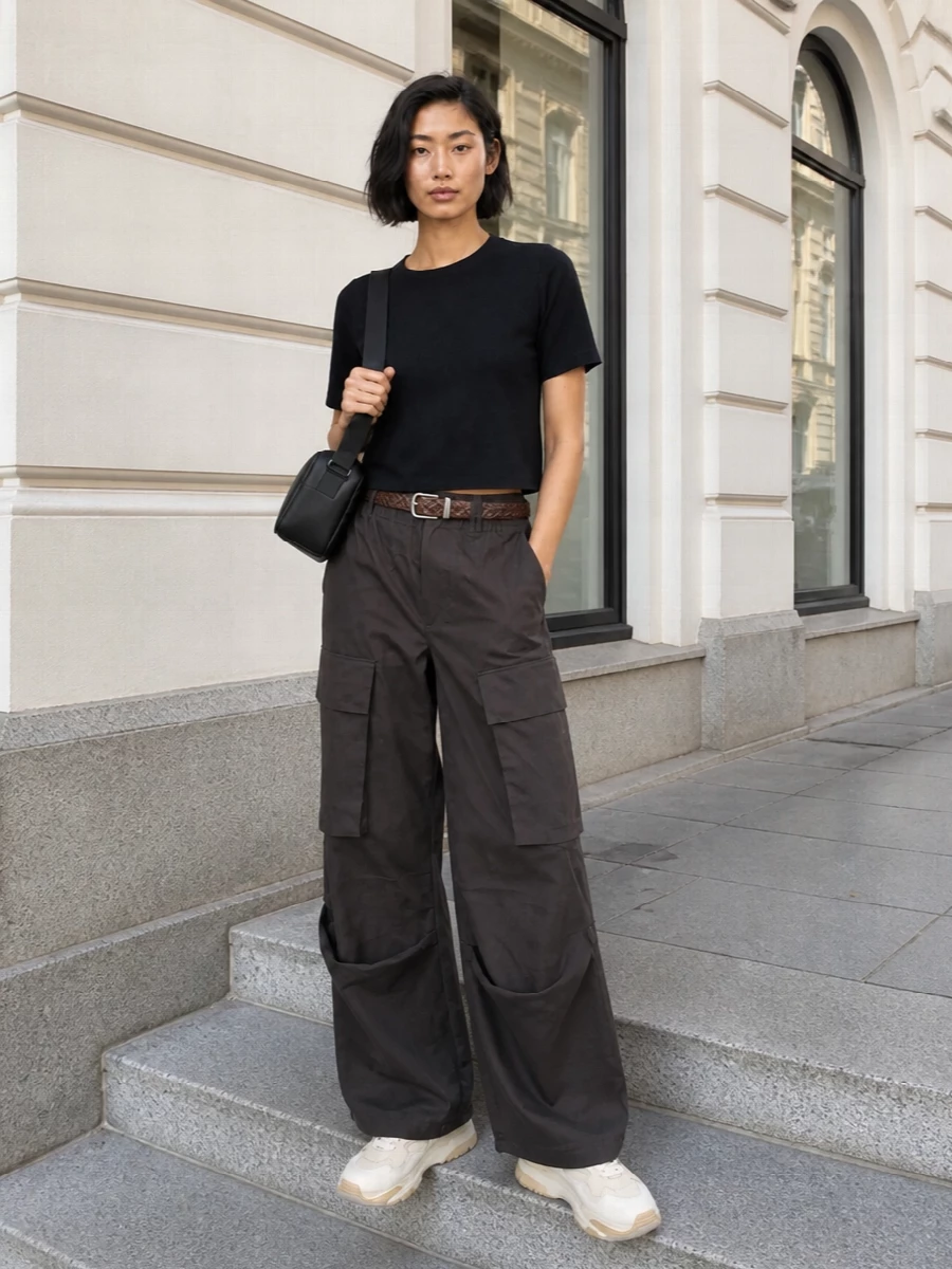 Charcoal cargos and the boxy black knit create a clean, utility-led base with a strong, modern line. Chunky neutral sneakers echo the volume at the hem, while the braided belt and compact cross-body keep the attitude practical yet refined for city days or travel. The palette stays grounded in deep browns and soft creams, so the whole outfit reads intentional rather than sporty.