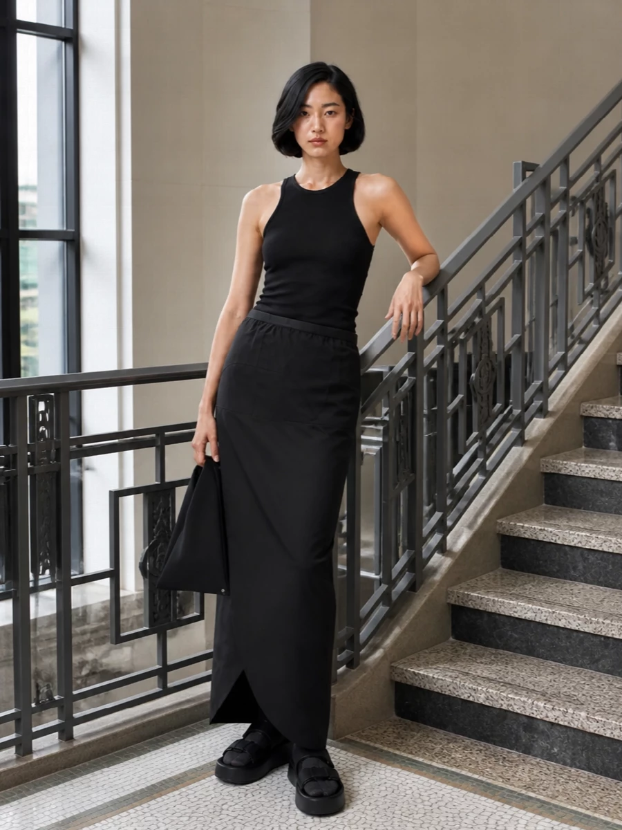 A black-on-black column that leans into Rick’s urban severity: the wrap maxi skirt grounded by a longline ribbed racerback tank. Chunky platform slides and a sculptural triangular tote keep the mood streetwise yet polished, with tabi socks adding a discreet, subversive detail at the base of the silhouette.