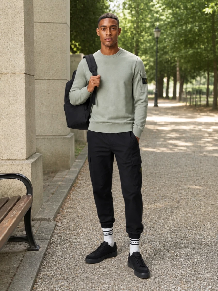 Stone Island cargo joggers ground the outfit in clean black utility, sharpened by a sage crew that adds just enough muted color. Black technical runners keep everything fast and streamlined, while the matching backpack and off-white sport socks nod to city movement without adding visual clutter. The result is a tight, technical street set that moves easily from commute to weekend.