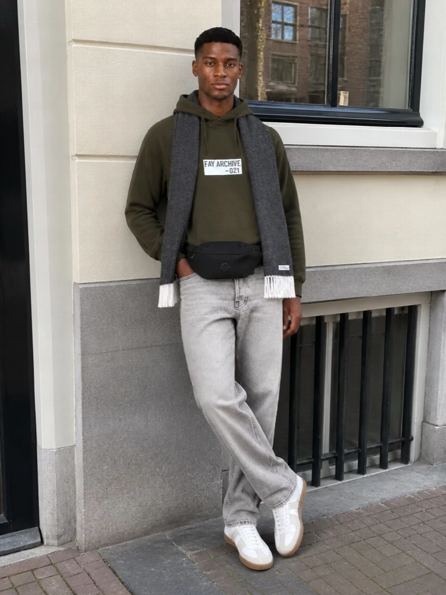 Olive and light-grey stay front and center, with the jean’s clean straight leg giving the hoodie a sharper, more intentional frame. White gum-sole sneakers keep it sporty yet minimal, while a black belt bag and charcoal scarf pull in darker notes for a balanced, city-ready finish. The result is relaxed streetwear that still feels considered from head to toe.