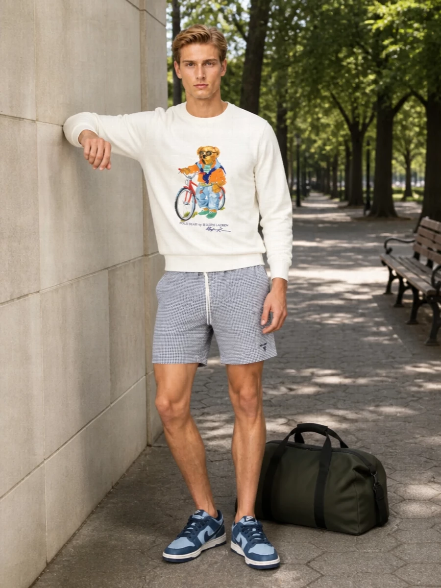 A preppy sport sweatshirt is paired with clean navy gingham shorts for a light, campus-adjacent take on summer streetwear. Navy and sky-blue sneakers tie into the graphic while keeping the palette tight and relaxed. A matte olive duffle finishes things with a practical, understated note that still feels ready for weekends on the move.