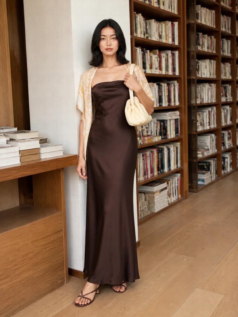 A liquid chocolate column is grounded with tonal leather sandals, then brightened by a buttercream clutch for a clean, modern evening mood. The silk foulard adds a whisper of soft gold at the shoulders, keeping the palette warm and romantic without breaking the minimalist line. It is an understated choice for summer dinners, rooftop drinks or a relaxed wedding guest moment.