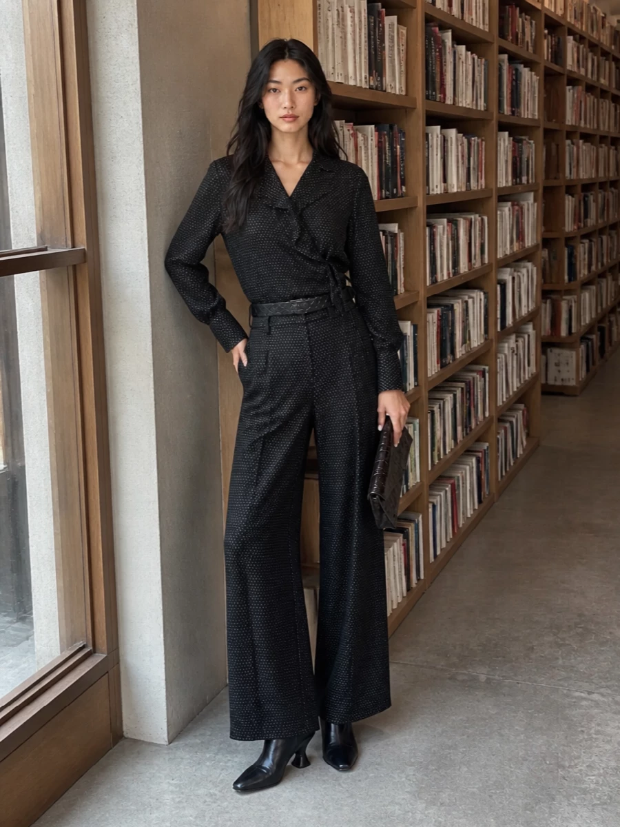 A studied Dries-on-Dries pairing turns the dotted wide-leg trouser into a modern, almost tuxedo-inspired ensemble, with the wrap blouse echoing its graphic yet restrained pattern. The sculpted black ankle boots and croc-embossed shoulder bag ground the inky palette, while a quiet black belt defines the waist so the volume reads deliberate rather than loose. It is polished enough for evening dinners, but still soft and languid in movement.