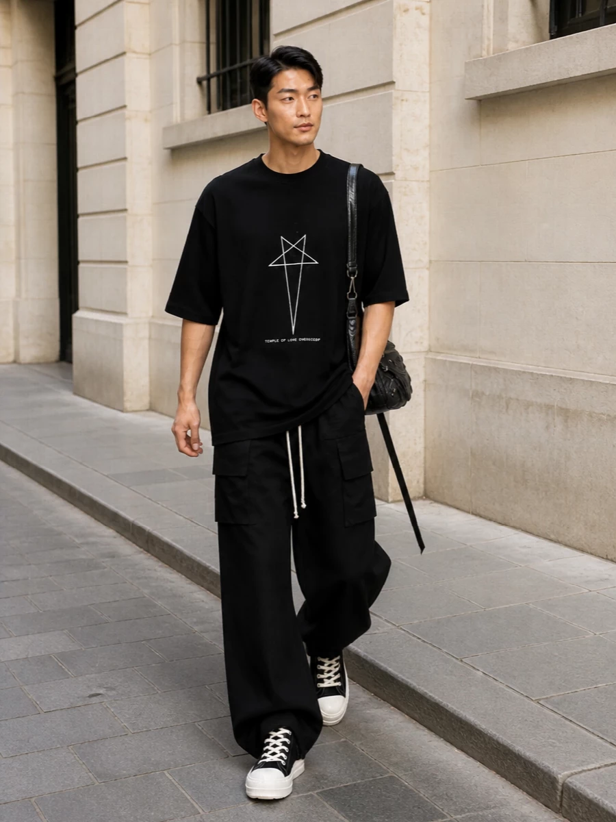 This look leans fully into Rick’s elongated, dystopian street uniform: the longline graphic tee drops over wide-leg black cargos, with off-white drawcords and graphic lines echoing across both pieces. Chunky black hi-tops with contrast soles ground the volume, while a compact, hardware‑rich shoulder bag adds a sharp, utilitarian finish that stays in step with the minimalist monochrome palette.