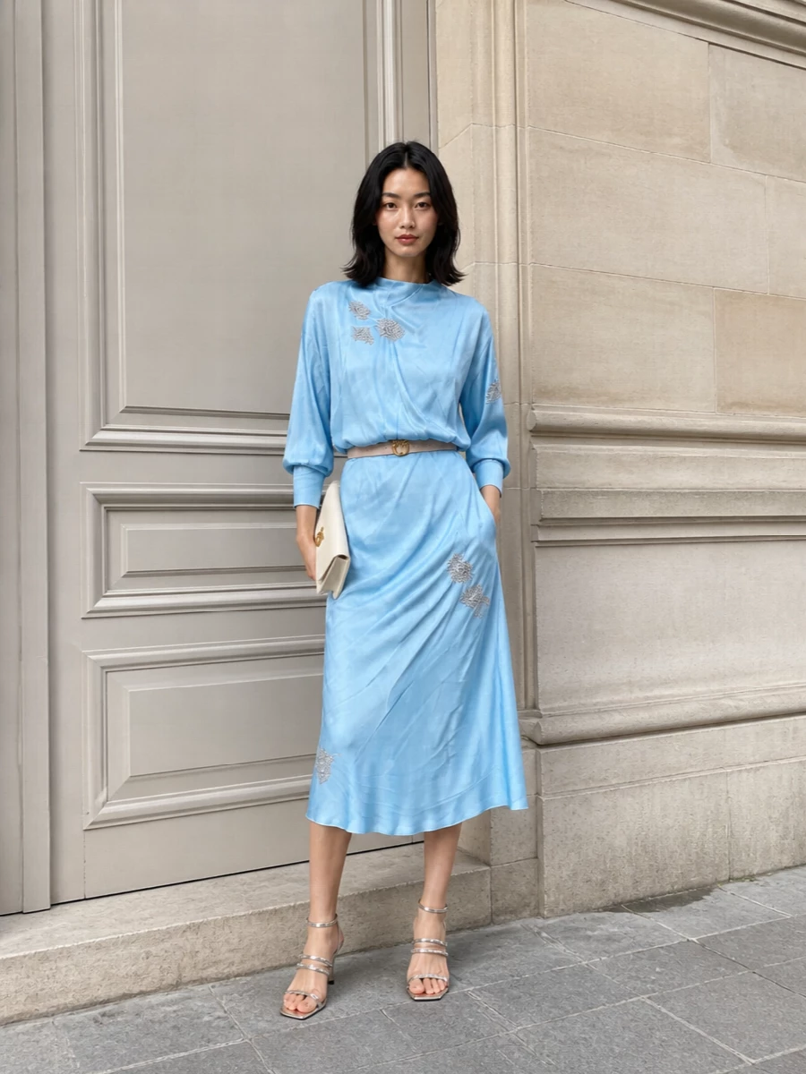 An ice-blue midi with subtle sparkle takes center stage, framed by silver straps and a clean ivory clutch for a soft, cocktail-ready palette. The pointed metallic heel keeps the mood romantic yet modern, while a slim blush belt quietly defines the waist without disturbing the dress’s drape.