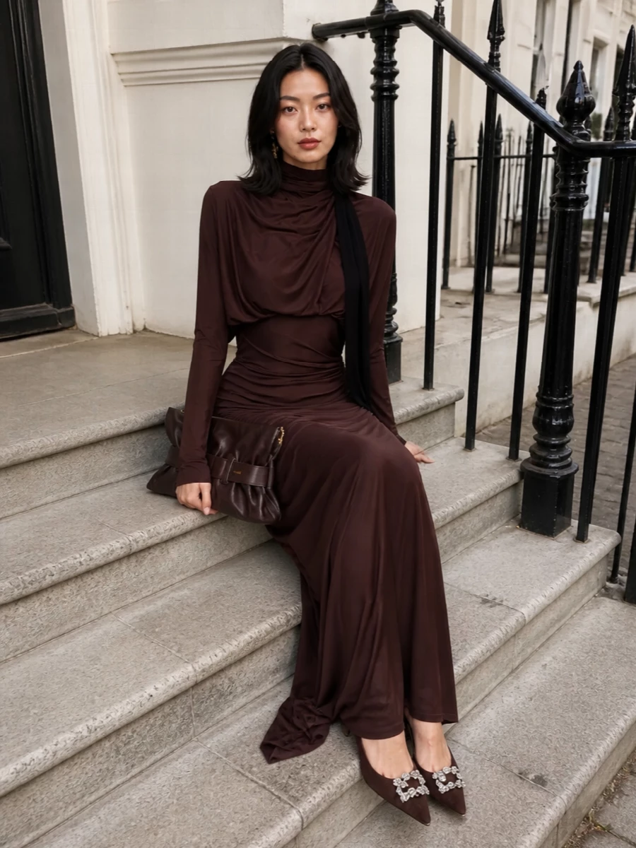 A deep chocolate Balmain gown pools around the body, its ruching and liquid jersey held in focus by tonal accessories that whisper rather than shout. Crystal-sparked pumps and a sculpted clutch carry the same rich brown story, while a single gold logo earring and a ribbon of black velvet at the neck add just enough light and structure. The result is sensual, high-formality eveningwear that feels elongated, composed and quietly powerful from every angle.