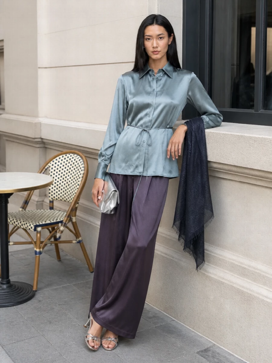Semicouture’s steel-blue satin shirt and matching plum wide-leg trouser read as a modern evening set, all fluid movement and softly defined shape. Silver strappy mules and a sculpted metallic clutch keep the palette cool and polished, adding just enough light without breaking the minimalist line. A gauzy navy wrap with a hint of sparkle finishes the outfit with discreet warmth and a whisper of shimmer at the shoulders.