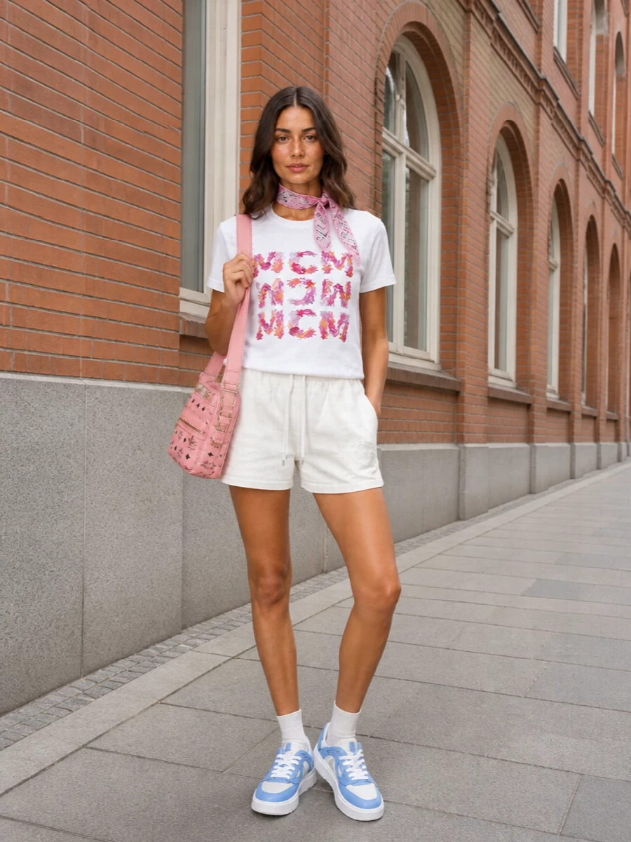 A white coral-logo tee pairs with clean soft-white shorts for an easy, leggy summer uniform that keeps the focus on colour and print. Chunky pale-blue sneakers ground the proportions while staying light, and a pink logo crossbody echoes the tee’s playful branding for a polished streetwear finish.
