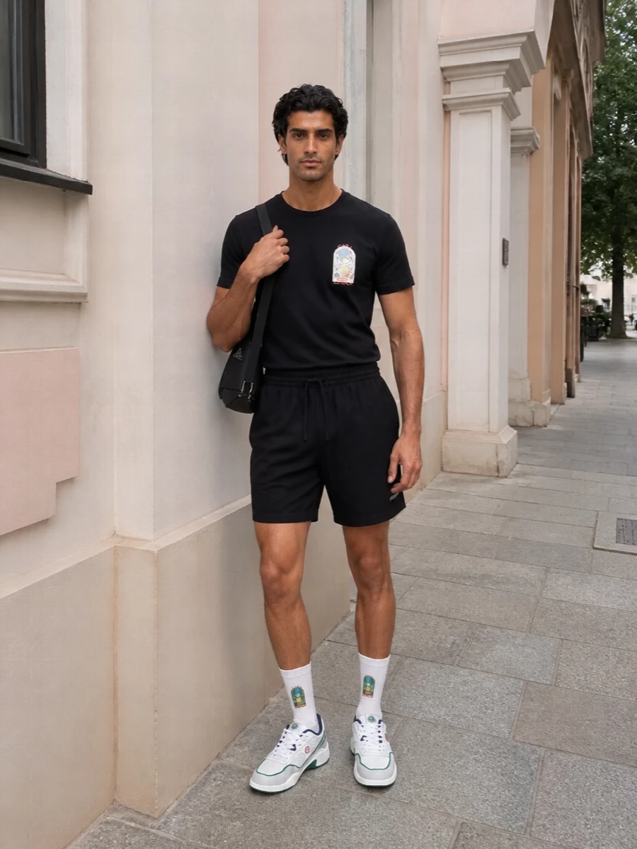 A black graphic tee and pared-back black deck shorts create a clean sport base that lets texture and proportion do the talking. White court sneakers and a sharp technical crossbody bring tennis-club polish into the city. Branded socks tie the story together with a subtle flash of color that stays within the resort-sport mood.