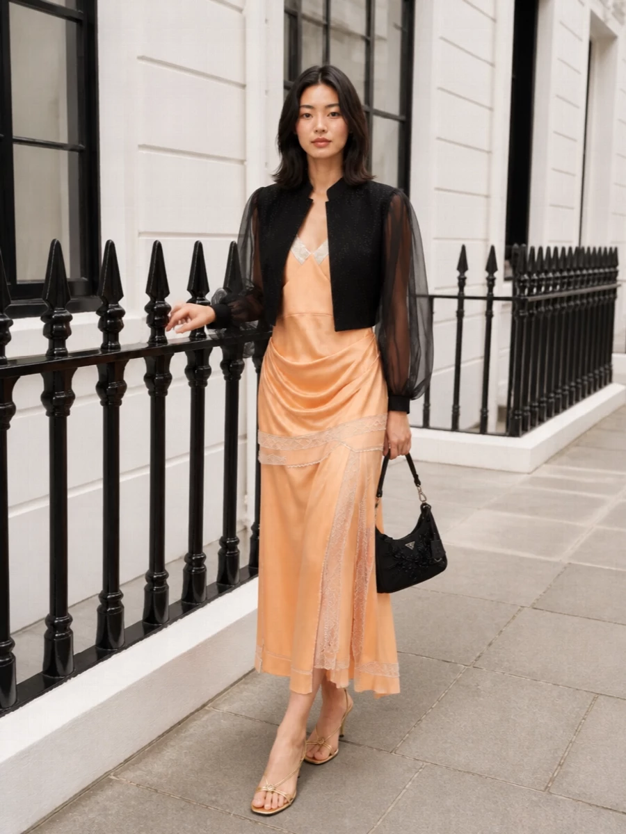 Magda Butrym’s peach satin slip stays centre stage, its draped lace-trimmed lines contrasted with a whisper of black and gold in the accessories for a refined, after-dark mood. The metallic sandal lengthens the leg and picks up the dress’s soft glow, while the beaded black baguette adds just enough texture and depth to keep the palette sophisticated rather than sweet. It is a romantic, quietly luxurious cocktail ensemble that moves beautifully from intimate dinners to late-night drinks in the city.