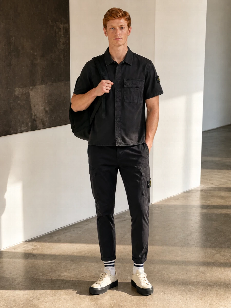 Head-to-toe Stone Island in deep charcoal and black keeps the cargo trouser feeling sharp rather than heavy, with the short sleeve shirt matching its utility lines. Neutral cream-toned sneakers and technical backpack lighten the mood without breaking the disciplined palette, so the whole outfit reads like a quietly functional city uniform. A hint of collegiate striping at the sock is the only contrast, giving the look some personality while staying restrained.