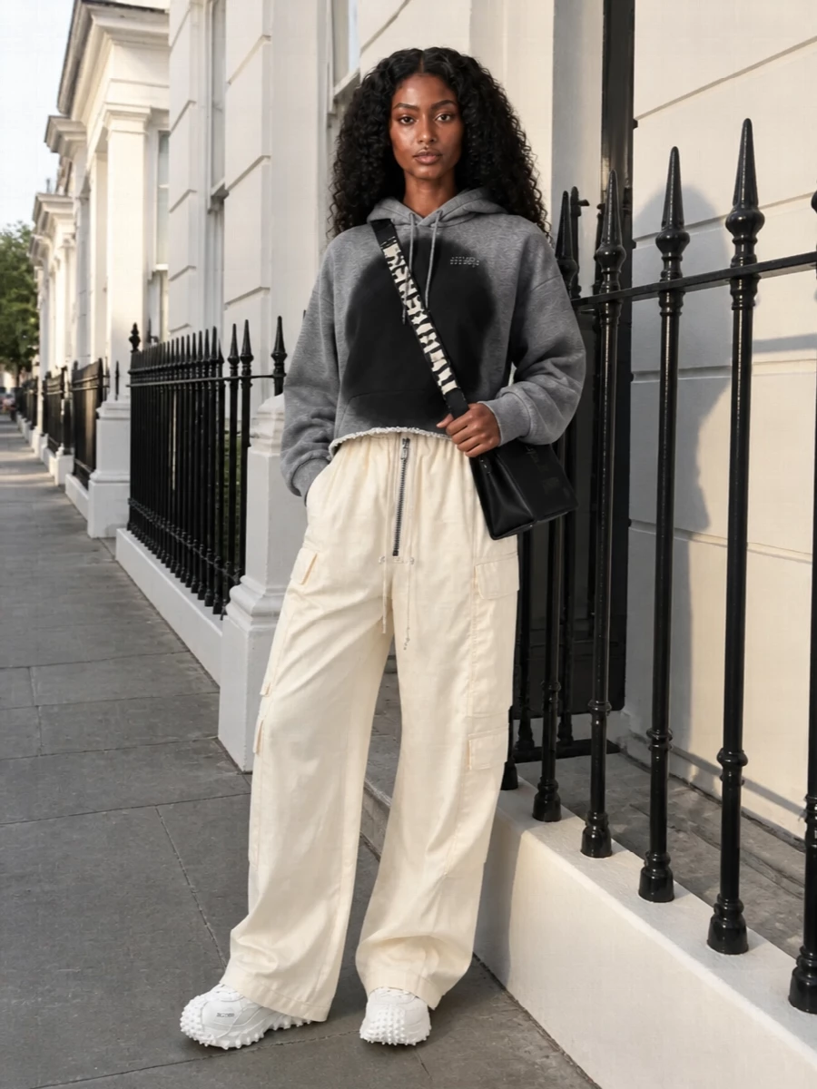 Chunky white Moncler sneakers ground a cropped sprayed Mm6 hoodie and wide Rick Owens cargos in a monochrome, techy street mood. The mix of sharp volume and neutral tones keeps it graphic but wearable for city days. A structured black crossbody adds just enough structure to all that movement without diluting the attitude.