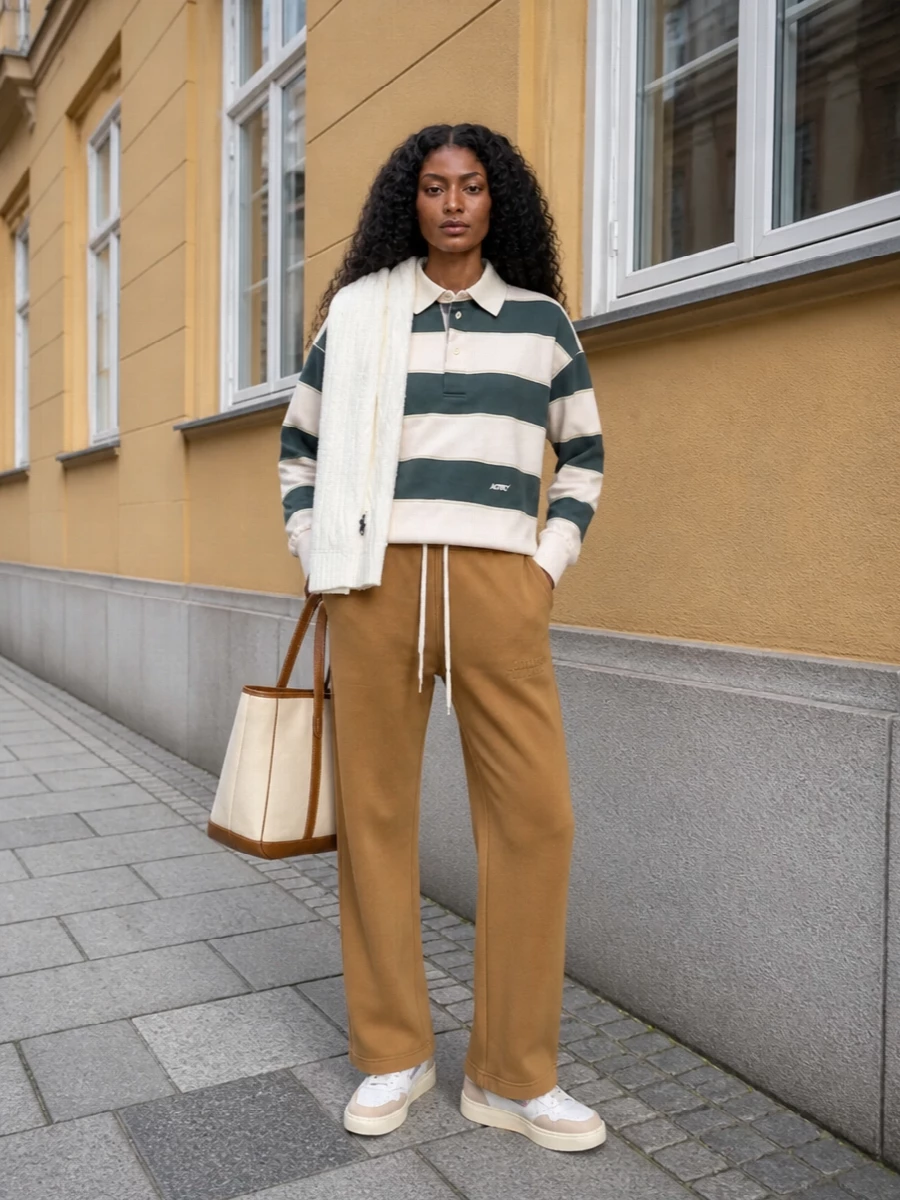 A relaxed rugby meets clean straight-leg track pants in warm camel for a collegiate, off-duty uniform that still feels intentional. Retro court sneakers and a structured canvas tote keep the palette soft and grounded, leaning into chalky neutrals with just enough contrast. The cable-knit scarf adds a final preppy note and a touch of winter brightness around the face.