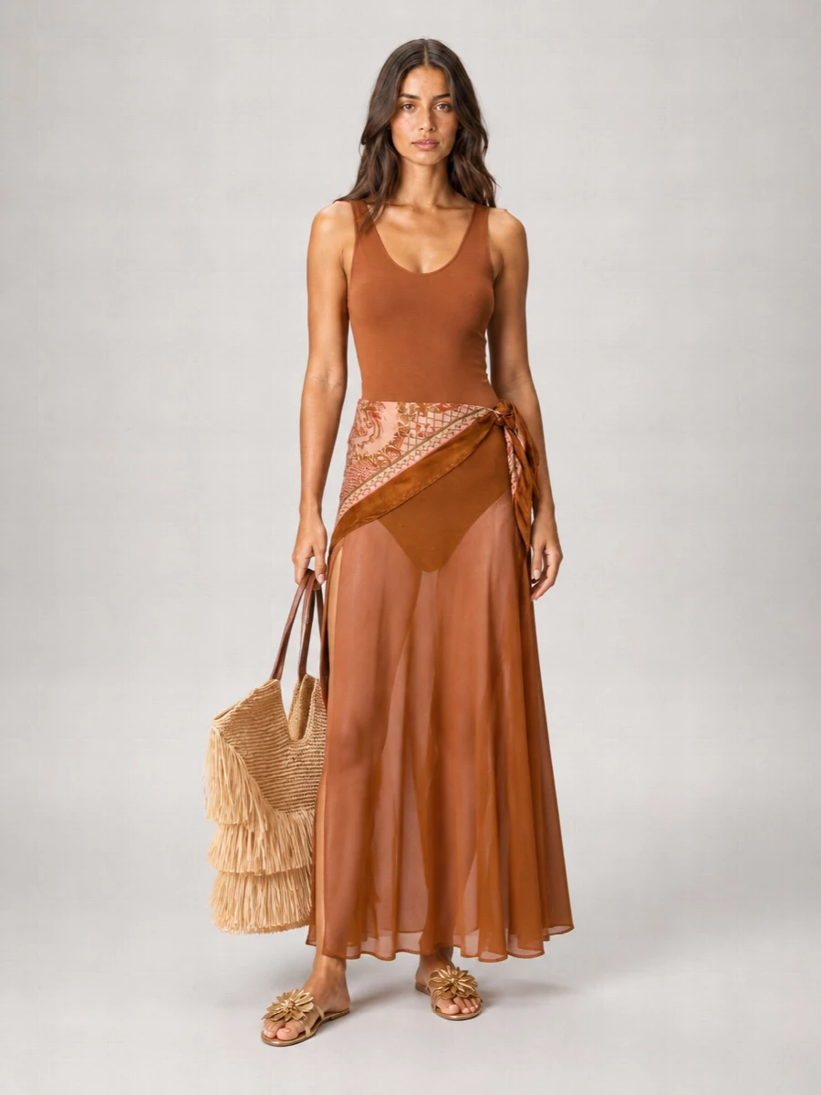 A sun-glazed take on resort romance, grounding the sheer rust maxi with metallic floral slides and a sweeping fringed straw tote. The palette stays in warm, toasted neutrals so the dress remains the quiet focus. Texture does the talking here, from gauzy layers to raffia fringe and petal-like leather, for an easy pool-to-cocktail transition.
