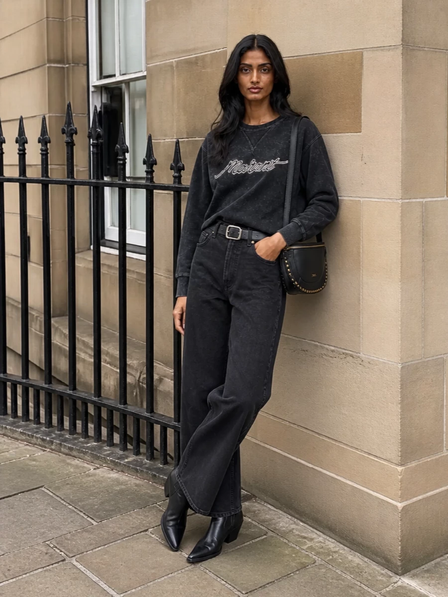 A washed-black Jean with a matching logo sweatshirt sets up a low-key monochrome base that feels easy but intentional. Ankle western boots sharpen the wide leg, while a compact studded crossbody and sleek belt keep the volume anchored and city ready. It is relaxed Parisian streetwear with just enough structure in the details.