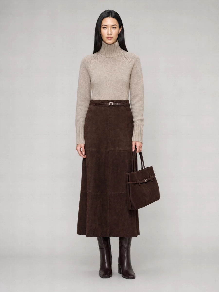 A high‑waisted chocolate suede midi skirt is softened with a taupe high‑neck knit, creating a calm play of depth and light within one tonal story. Knee‑high chocolate boots and a structured suede tote echo the skirt’s richness, while a slim dark‑brown belt quietly defines the waist for a long, composed line. The result feels quietly assured and modern, ideal for city days that need polish without noise.