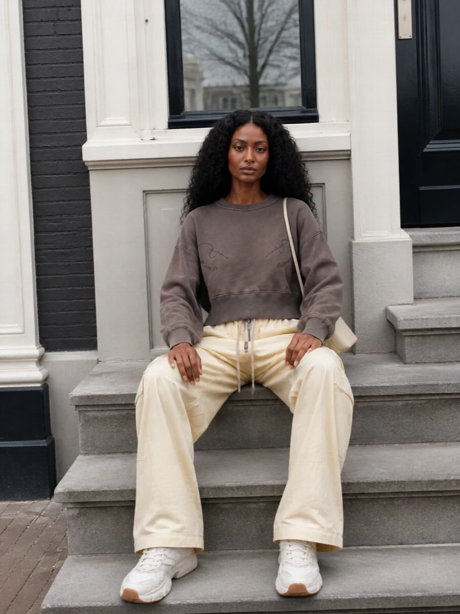 Soft neutrals and easy volume keep this outfit grounded in modern streetwear, with the chunky runner echoed by wide buttercream cargos and a cropped taupe sweatshirt. The proportions stay relaxed but intentional, with the waist definition and long leg line preventing the pieces from feeling sloppy. A slim buttercream cross-body finishes it with just enough structure to move from coffee runs to low-key dinners in one go.