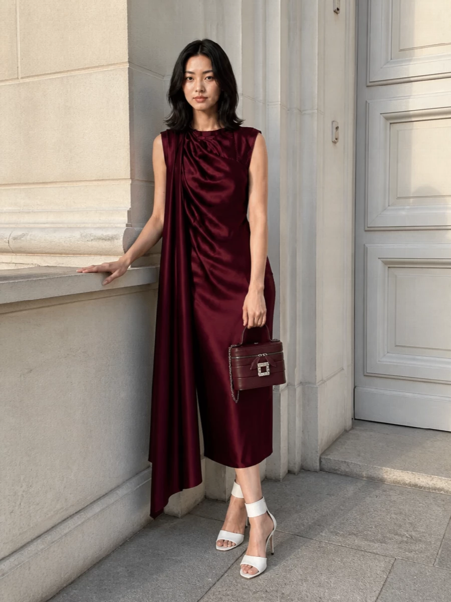 A deep wine satin column against crisp white leather heels creates a precise, romantic evening statement that feels modern rather than overt. The asymmetric drape of the dress picks up the sculpted line of the sandal, while the burgundy box clutch quietly reinforces the palette instead of competing with it. Choosing the dress route over separates keeps the silhouette clean and uninterrupted, which best serves the shoes’ couture ankle cuff and the quiet luxury mood.