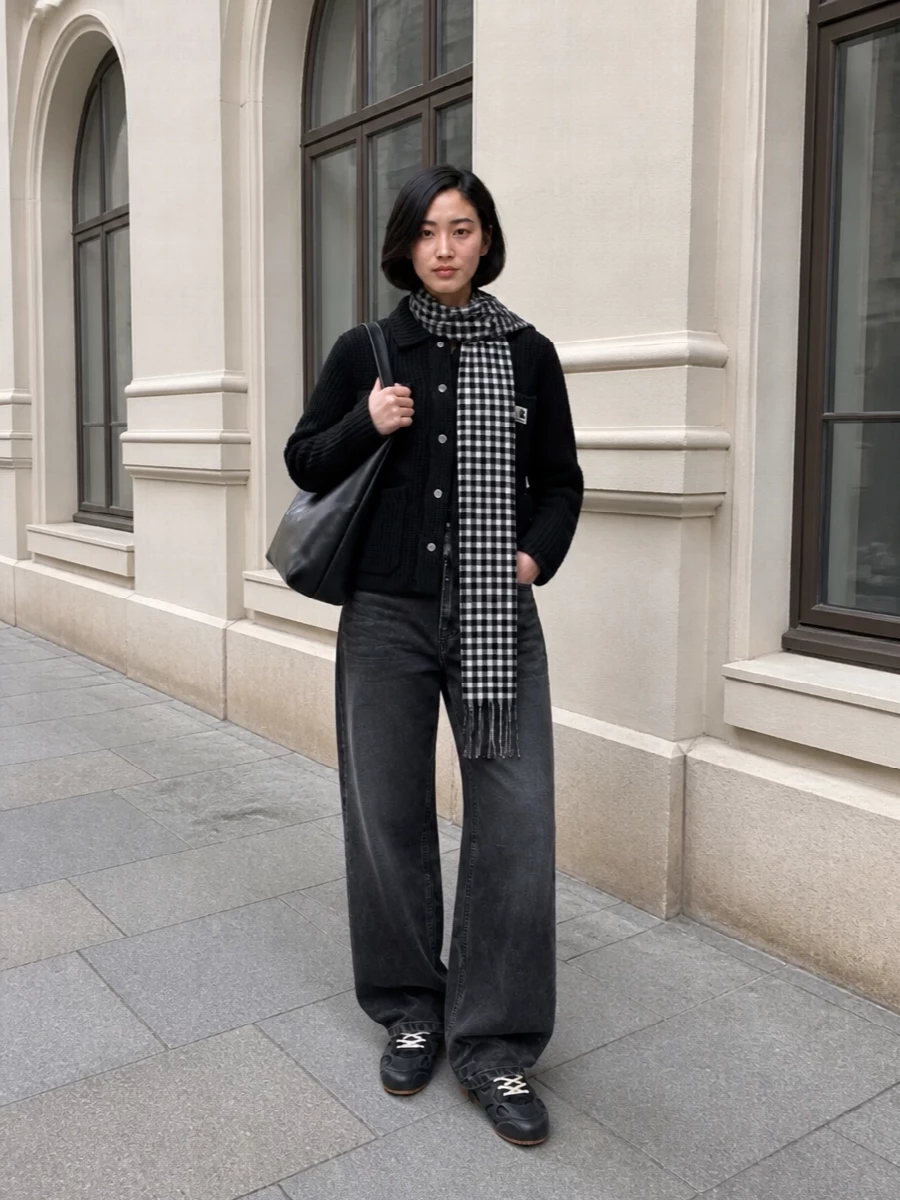 A boxy black knit and charcoal wide-leg denim keep the silhouette strong and grounded, with workwear details reading quietly utilitarian rather than heavy. Dark sneakers and a soft black shoulder bag lean it into the city, while the monochrome scarf adds a sharp graphic note without disturbing the minimal palette.