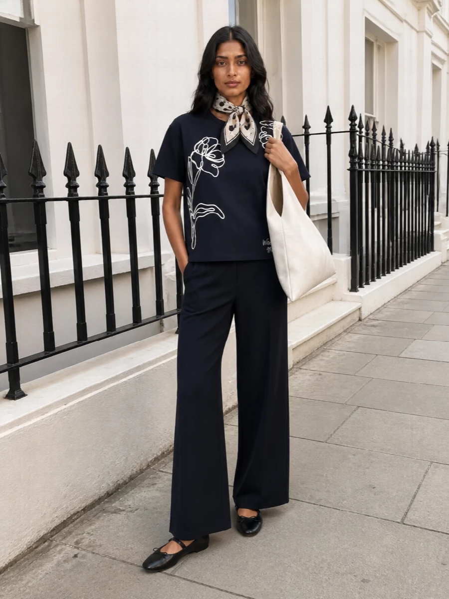 Navy-on-navy keeps this outfit calm and composed, letting the sketched florals and wide-leg volume do the talking. The soft, fluid trouser elongates the line under the boxy tee, while black Mary Jane flats and an ivory origami tote sharpen the finish just enough for city days. A heart-print scarf at the neck or bag adds a gentle romantic note that still feels clean and modern.