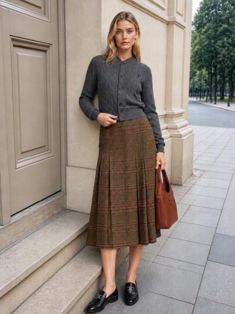 Charcoal cable knit meets heritage plaid for a quietly polished, preppy take on cold-weather dressing. The high-waisted Saint Laurent midi defines the waist and lengthens the line, while sleek Gucci loafers keep the mood collegiate rather than corporate. A structured Miu Miu top-handle in rich chestnut finishes the palette with warmth that feels considered from office hours to city weekends.